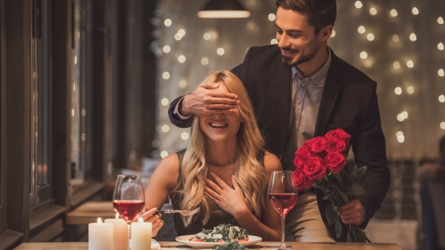 Handsome elegant man is holding roses and covering his girlfriend's eyes while making a surprise in restaurant, both are smiling