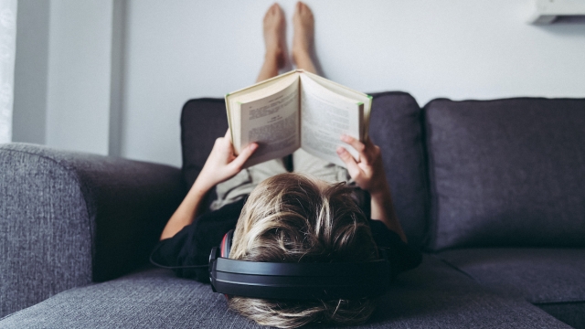 Blond boy barefoot reads book and listening music lying down. Student reading the lesson comfortably relaxed, diligent child reads a story young man plunges into culture Home works and relax on sofà
