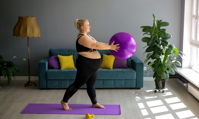 A fat, obese pregnant lady is doing Pilates at home, lifting a fit ball above herself. Home workouts in Interior room with a large window, sofa and yellow-purple pillows. Weight loss in quarantine