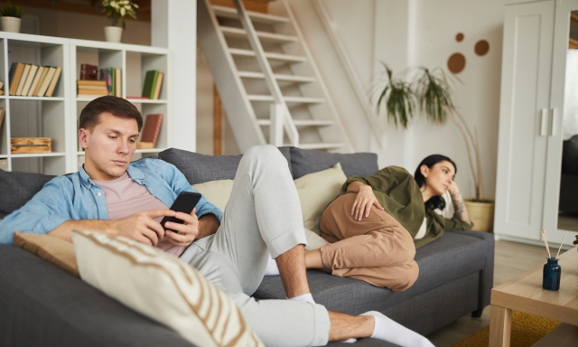 Full length portrait of bored young couple watching TV on sofa at home, focus on man using smartphone in foreground, copy space