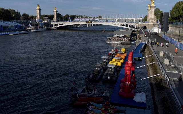 Watercraft and buoys sit along the Seine river as the triathlon event venue on the Pont Alexandre III bridge stands in the background at the 2024 Summer Olympics, Sunday, July 28, 2024, in Paris. (AP Photo/David Goldman)