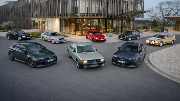 Several Audi RS models from different generations with five-cylinder engine are arranged in a semicircle in front of a modern glass building.