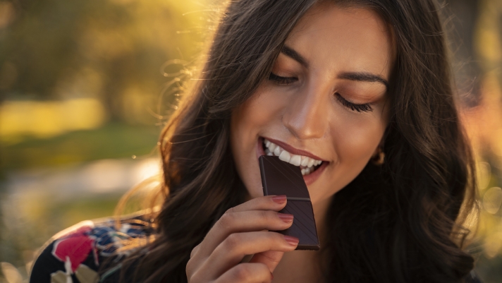 Natural brunette woman taking a bite of organic dark chocolate outdoors on sunny day