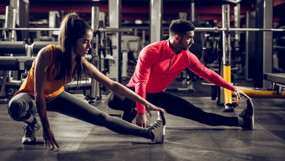 Young healthy sporty active shape girl with a ponytail doing leg stretches on the floor while crouching with a handsome helpful personal trainer next to her in the gym.