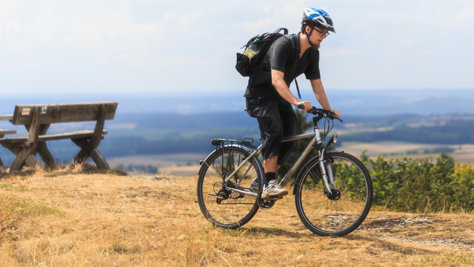 Biking in the Franconian Hills in Northern Bavaria. Caucasian Young man in black sports outfit with his Trekking bike on a rock, looking into the distance. Shot in High Summer August on an afternoon