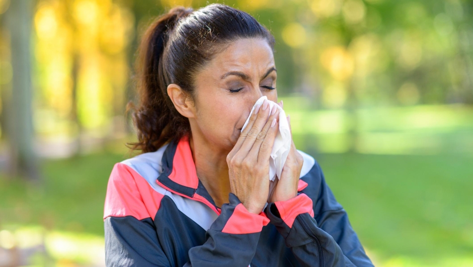 Woman blowing her nose on a tissue outdoors in a leafy green park while out jogging conceptual of seasonal flu or allergies