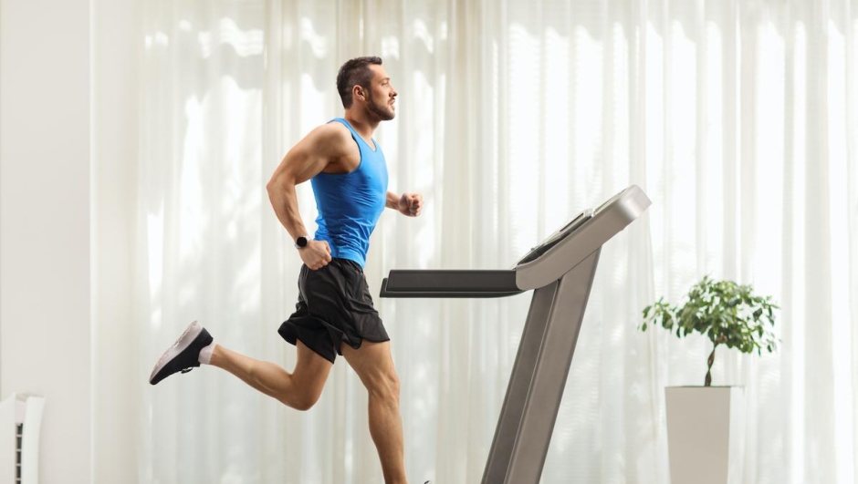 Full length profile shot of a young man running on a treadmill at home