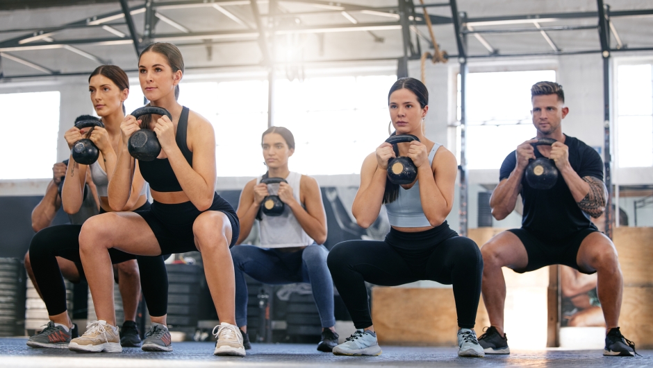Diverse group of active young people doing kettlebell squat exercises while training together in a gym. Focused athletes challenging themselves by holding heavy weights to build muscle and endurance during a workout in a fitness class