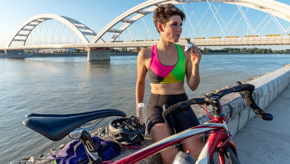 Happy young woman cyclist eating an energy bar and resting by the river bank, after riding a bicycle. Healthy lifestyle and sports concept.