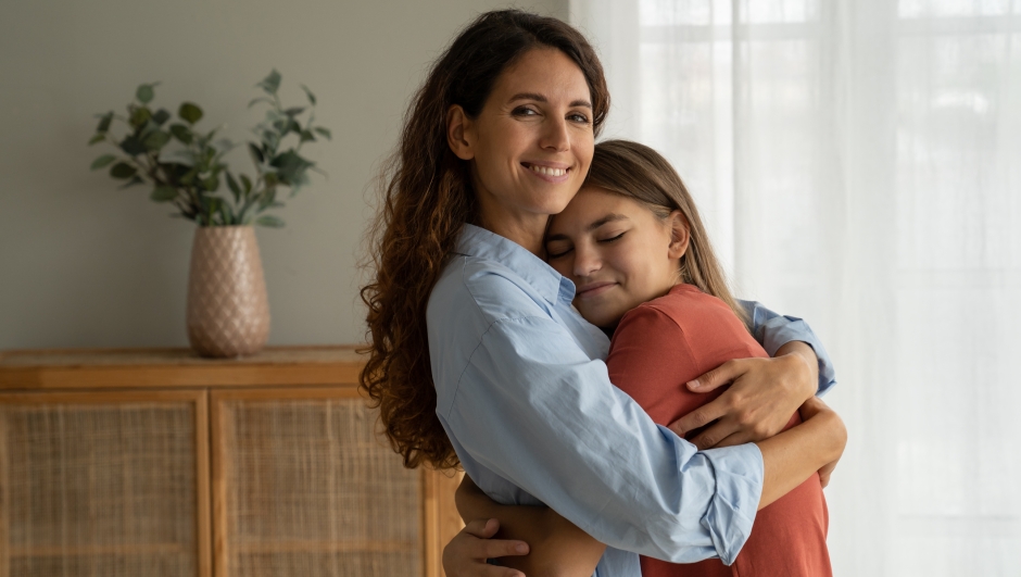 Young loving mother hugging cuddling with teen girl daughter at home and smiling at camera, expressing unconditional love. Mother-daughter friendship, happy motherhood, parenting of adolescent