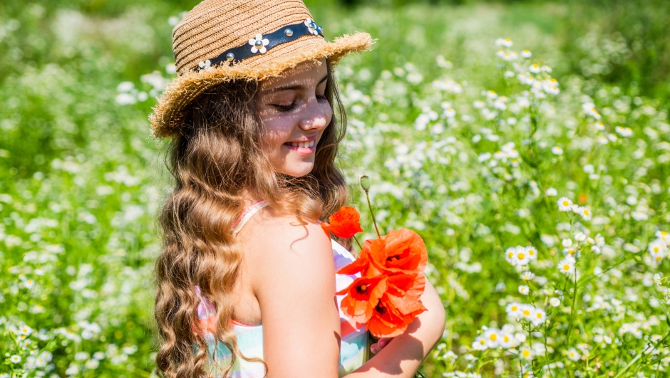 Happy small girl with beauty look wear sun hat on long curly blonde hair holding poppy flowers on natural sunny summer landscape, salon.