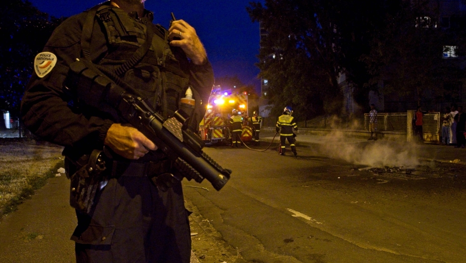 A police officer holds a flash ball as he stands next to firefighters during clashes on June 30, 2023 in Tours, France, three days after a 17-year-old man was killed by police in Nanterre, a western suburb of Paris. France on June 30 deployed 45,000 officers backed by light armoured vehicles to tackle a fourth straight night of violent protests after the fatal police shooting of a teenager. Crack police units and other security forces fanned out across the country to quell violence over the shooting, which took place during a traffic stop in a Paris suburb on June 27. (Photo by GUILLAUME SOUVANT / AFP)