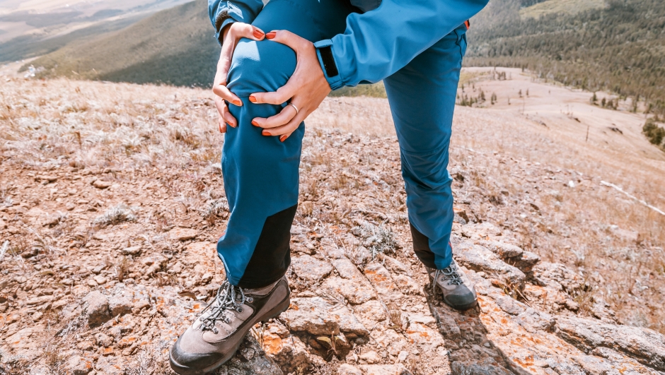 Woman is holding on to the knee joint, suffering from acute pain. Concept of injury of meniscus during a hike in the highlands