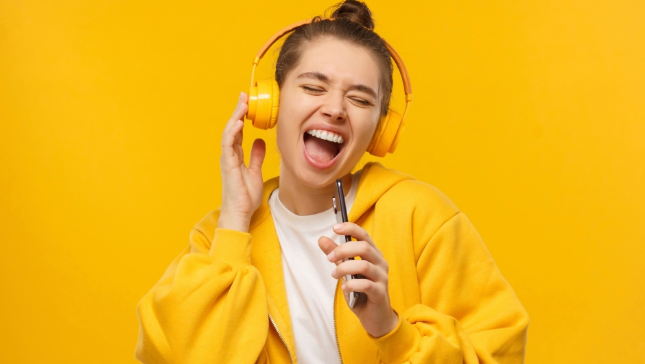 Trendy girl singing favorite song out loud in phone as mic, wearing wireless headphones, isolated on yellow background. Karaoke online app.