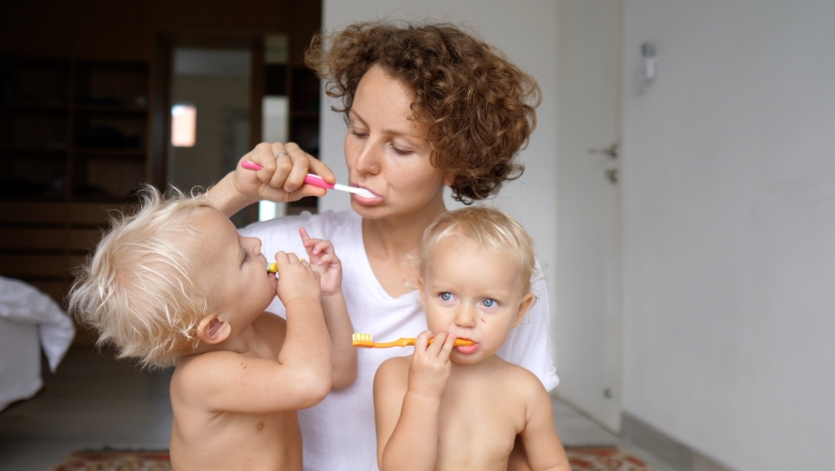 A young mother teaches her twins how to brush their teeth by example. She clearly shows the children what manipulations need to be carried out. Hygiene rituals as a time of quality communication.