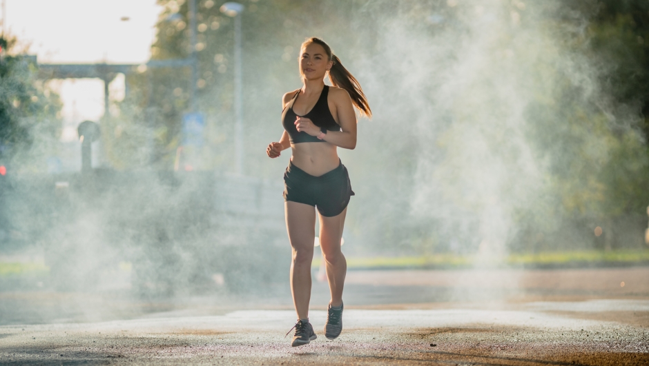 Beautiful Fitness Girl in Black Athletic Top and Shorts is Energetically Running in the Street. She is Jogging in an Urban Environment with Blurred Cars in the Background.
