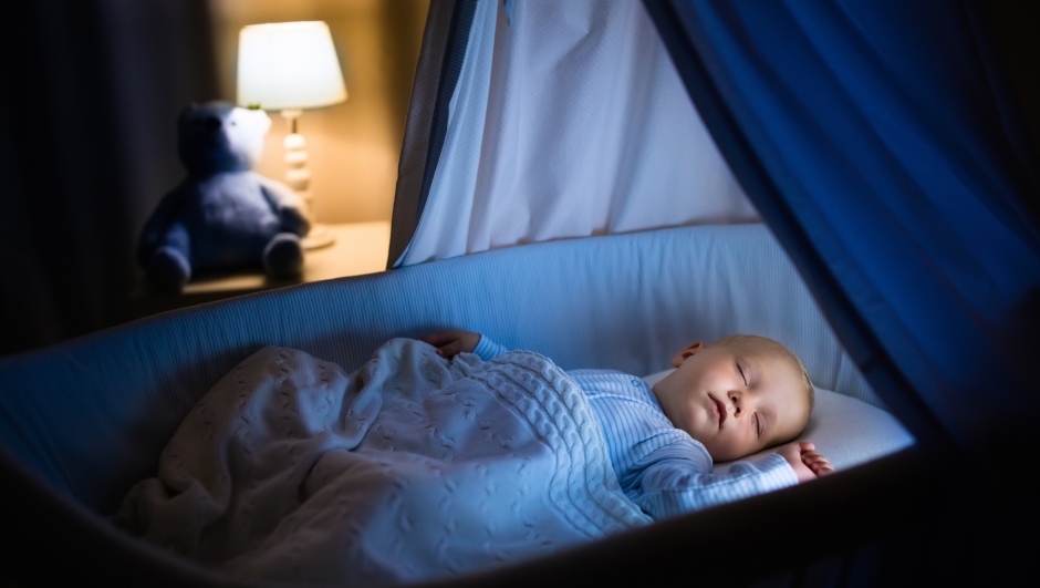 Adorable baby sleeping in blue bassinet with canopy at night. Little boy in pajamas taking a nap in dark room with crib, lamp and toy bear. Bed time for kids. Bedroom and nursery interior.