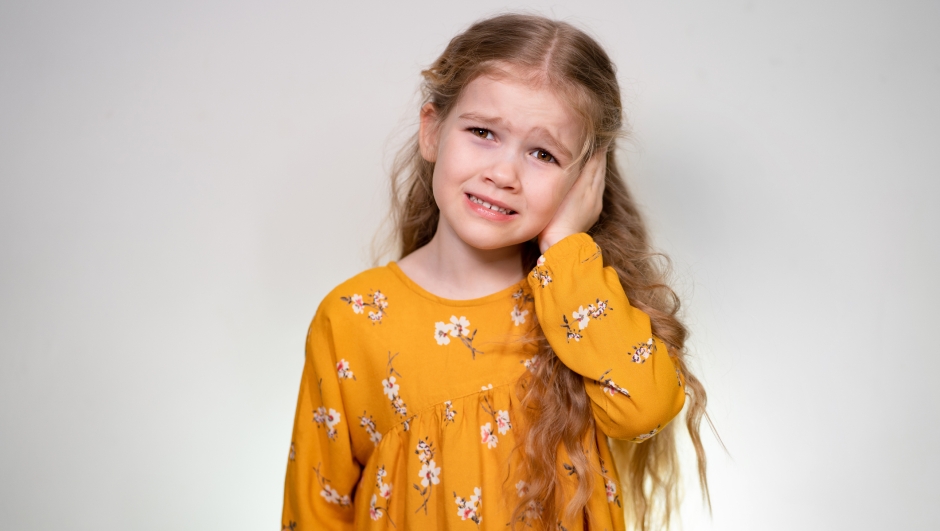 A little girl's sore ear and she holds his hand and cries. Babe posing in yellow dress on the white background of the wall.