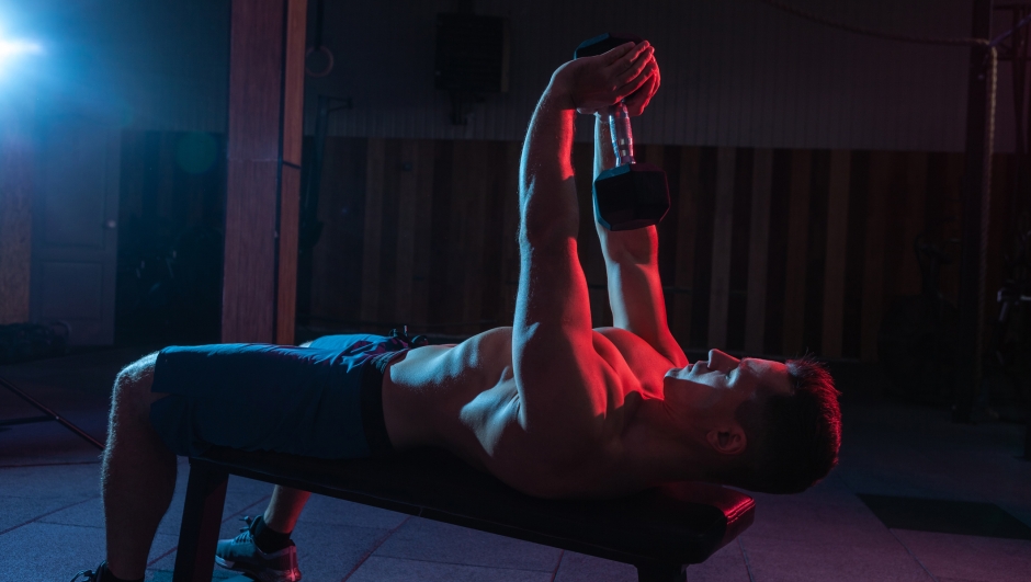 Muscular man trains with dumbbell in red-blue gradient light in dark cross fit hall