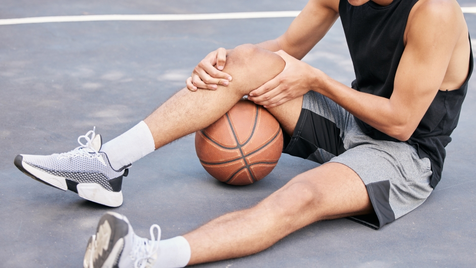 Basketball, man and knee in sports injury on the court holding painful, sore or tender area in the outdoors. Basketball player suffering from leg pain, joint or inflammation in sport match or game