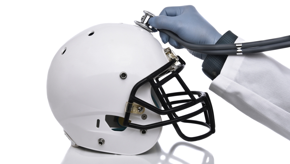 A football helmet and doctors hand holding a stethoscope on the crown of the helmet. Sports Concussion Concept, and related conditions, CTE, Alzheimer's, Parkinson's.
