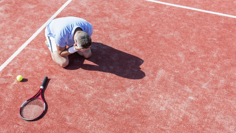 High angle view of disappointed mature man with head in hands while kneeling by tennis racket on red court during summer