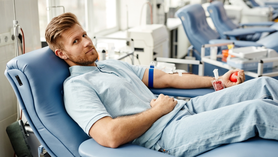 Full length portrait of young man donating blood while lying in comfortable chair at med center and looking at camera