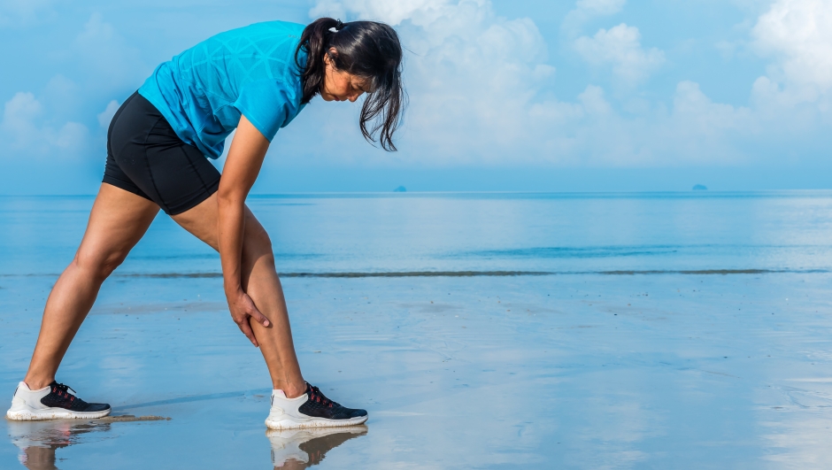 Woman has calf cramp pain of her legs during outdoor running at beach. Calf muscle jogging injury, healthy lifestyle outdoor exercise concept.