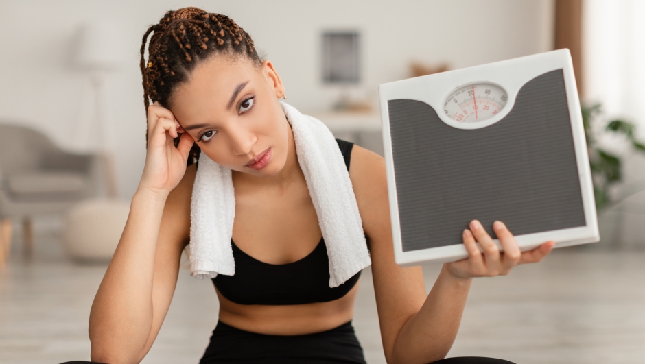 Weight Loss Failure. Unhappy Fitness Woman Showing Scales After Unsuccessful Slimming Looking At Camera Sitting At Home, Wearing Sportswear. Weight Gain Issue Concept