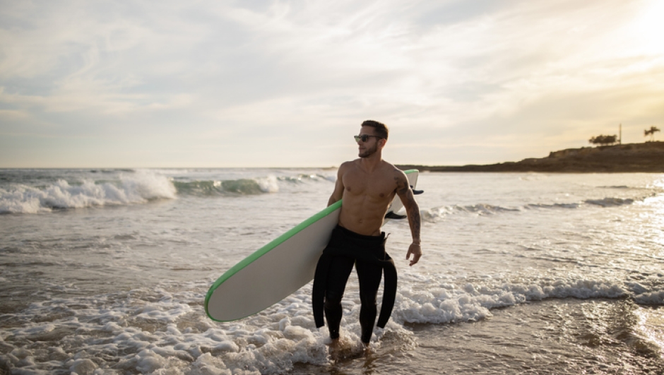Surfer Guy. Young Man In Wetsuit Carrying Surfboard While Walking On Seashore, Sporty Athletic Male Ready To Ride Waves, Enjoying Surfing At Sunset, Handsome Man Doing Water Sports, Copy Space