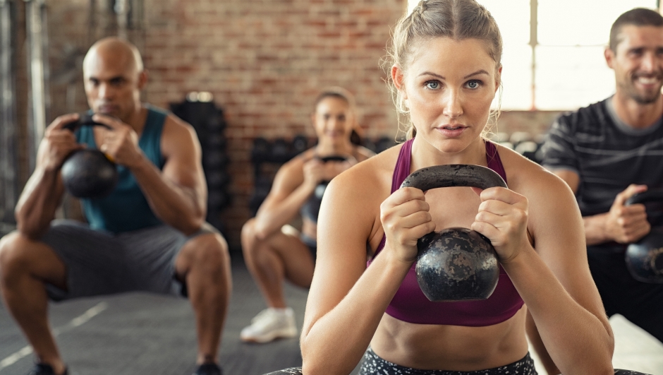 Group of fit people holding kettle bell during squatting exercise at crossfit gym. Fitness girl and men lifting kettlebell during strength training exercising. Group of young people doing squat with kettle bell.