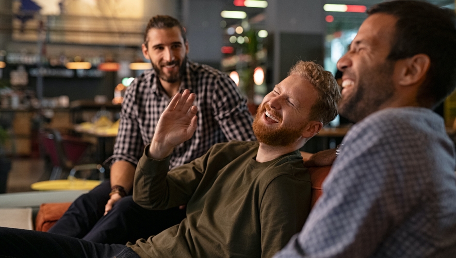 Group of three best friends laughing and enjoying the evening at pub. Happy young men enjoying late night staying together at bar. Cheerful guys sitting on couch and having fun while relaxing after work.