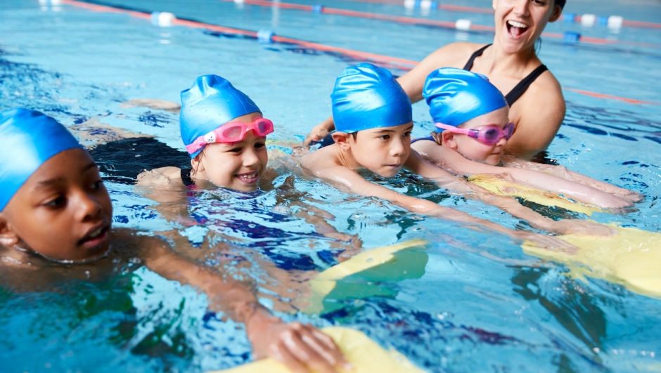 Female Coach In Water Giving Group Of Children Swimming Lesson In Indoor Pool