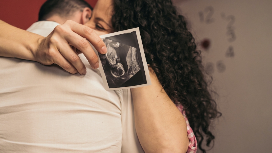 Pregnant Woman Embracing Future Baby’s Father Holding Printed Ultrasound. Young pregnant girl hugging partner holding baby’s ultrasound print.