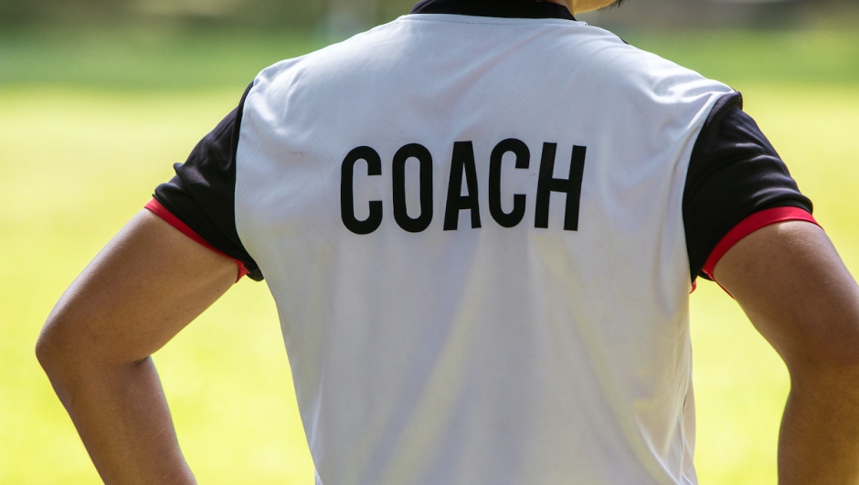 Back view of male soccer or football coach in white shirt with word COACH written on back, standing on the sideline watching his team play, good for sport or coaching concept