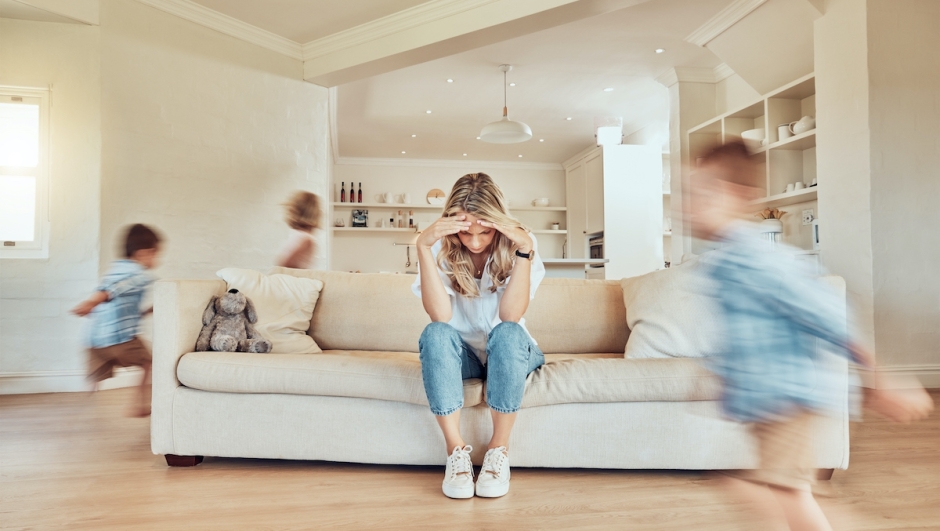 Stressed mother sitting on the sofa while her adhd children run around the living room. Hyperactive son and daughter giving single parent a headache. Active siblings chasing each other in the house