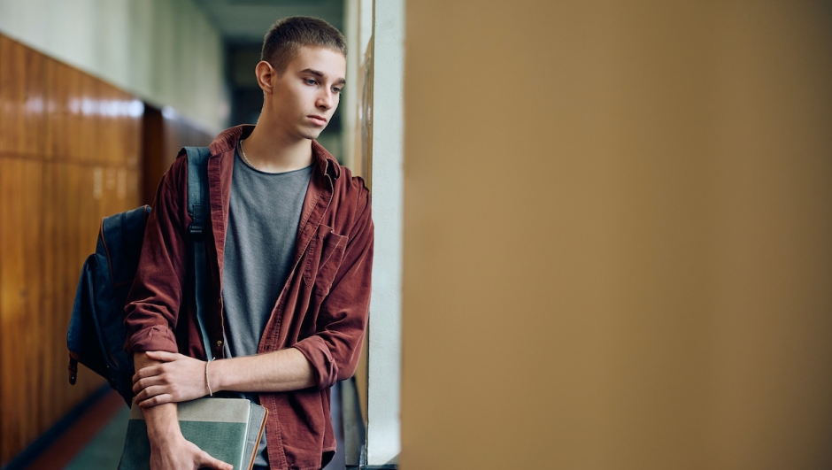 Sad teenage boy thinking of something while standing in hallway at high school. Copy space.