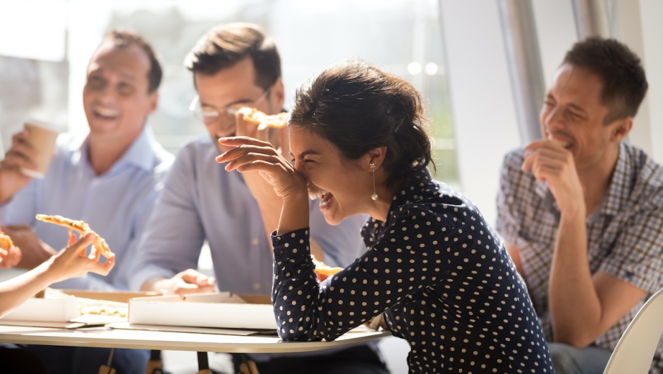 Indian woman laughing at funny joke eating pizza with diverse coworkers in office, friendly work team enjoying positive emotions and lunch together, happy colleagues staff group having fun at break