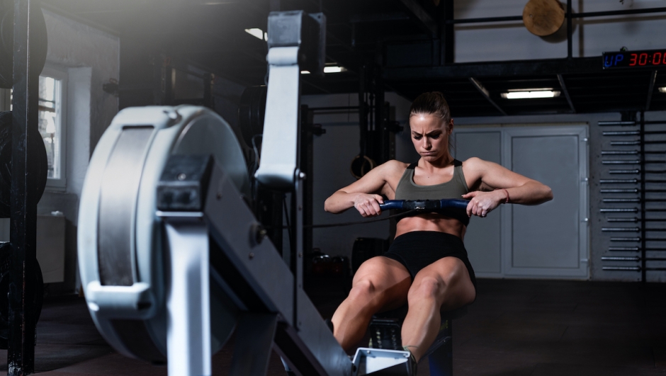 Young strong fit sweaty powerful attractive muscular woman with big muscles doing hard core row heavy cross training workout on indoor rower at the gym real people exercise