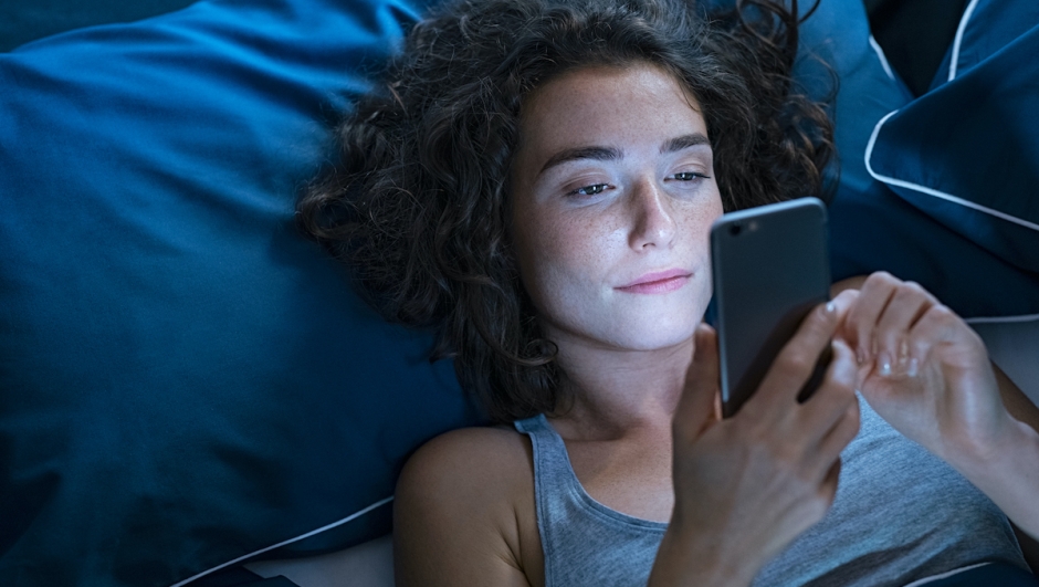 Top view of young woman using a smartphone while lying on bed in the night. High angle view of pretty girl messaging on smart phone before sleeping at night. Addicted woman suffering from insomnia and chatting and surfing on the internet with her cellphone late in night.