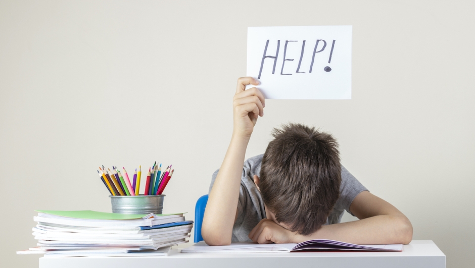 Sad tired frustrated boy sitting at the table with many books and holding help sign. Learning difficulties, education concept.