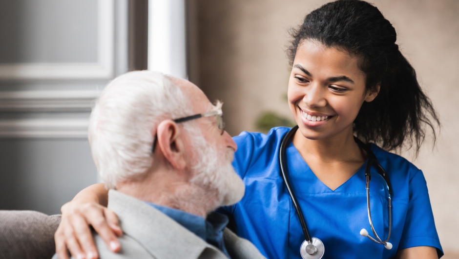 Caregiver supporting happy disabled older man sitting close up, touching shoulders, expressing care and love, smiling nurse wearing blue uniform and mature patient having fun