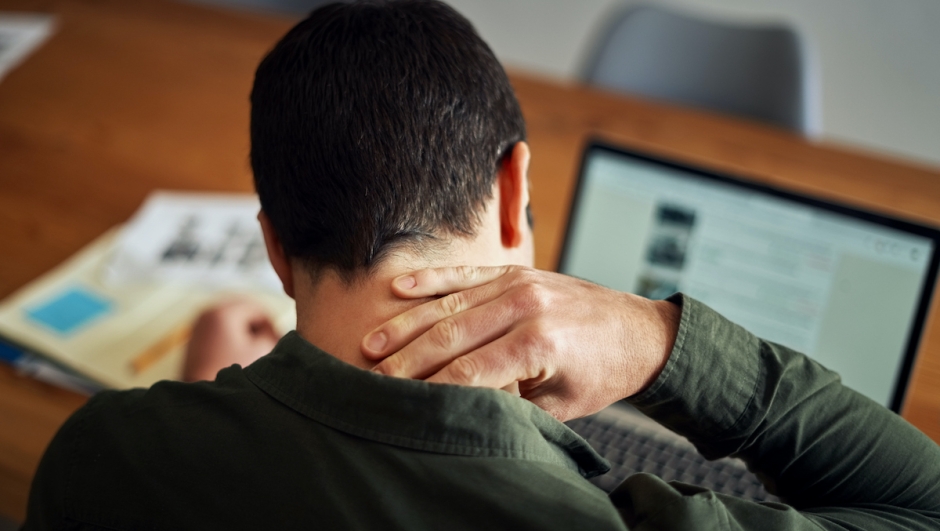 Rear view of a man suffering from neck pain while working on laptop in table