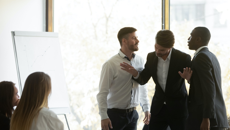 Millennial businessman setting apart two aggressive multiracial coworkers, starting fighting at office. Mixed race young colleagues quarreling shouting at each other, misbehaving during meeting.