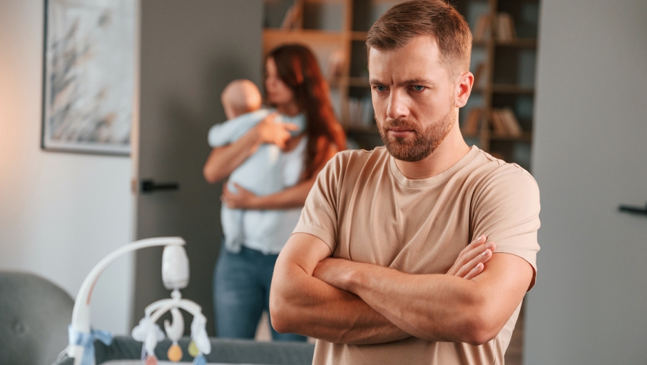 Offended man standing with arms crossed. Couple with newborn baby is at home having conflict.