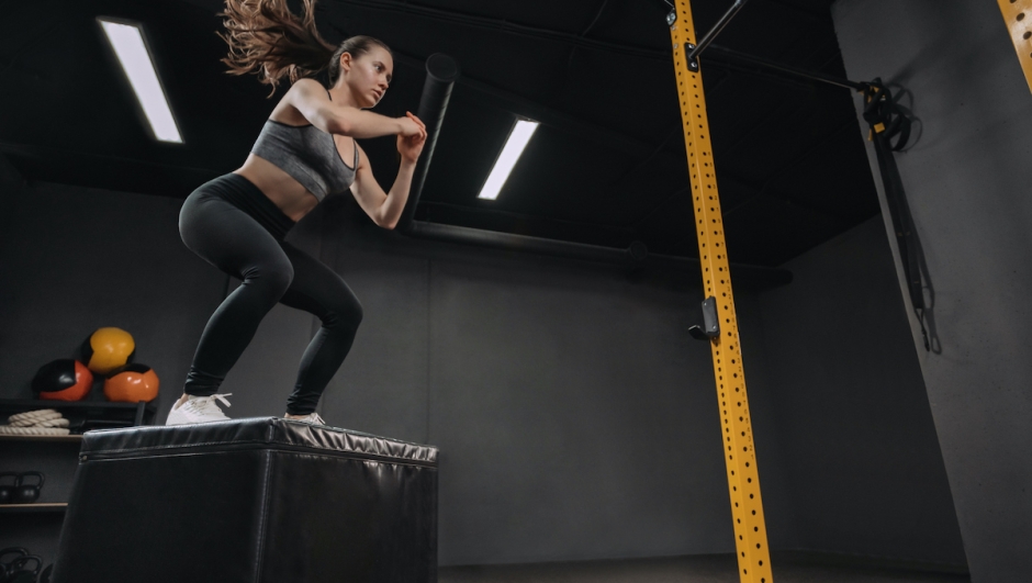 Woman doing box jump exercise as part of her crossfit training. Female athlete doing squats and jumping onto the box in dark workout gym. Copy space