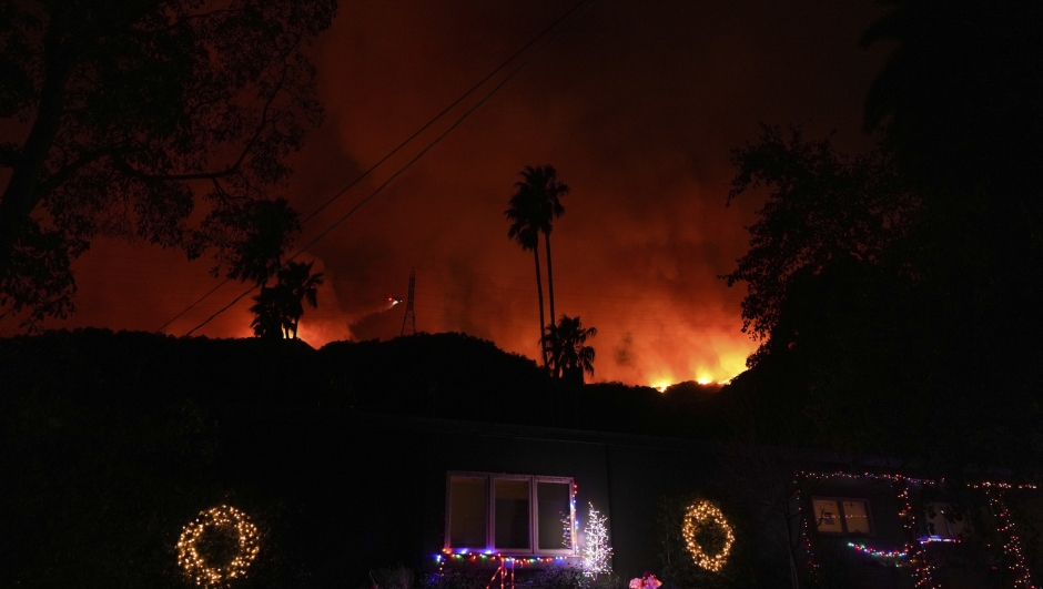 A helicopter drops water on the Palisades Fire behind homes decorated with Christmas lights in Mandeville Canyon on Friday, Jan. 10, 2025, in Los Angeles. (AP Photo/Eric Thayer)