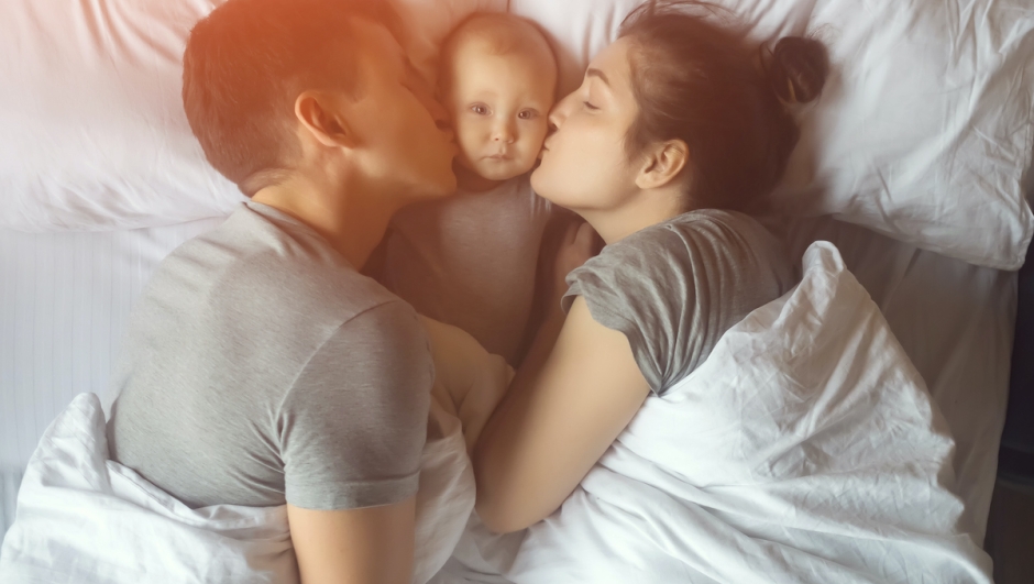 Young mother and father lies on bed with baby girl in early morning, sunlight. Parents kiss little child on cheek, top view.