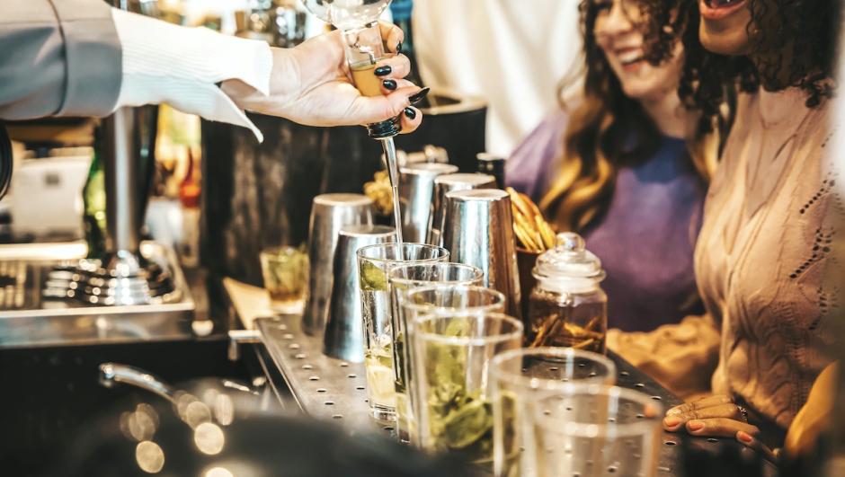 Bartender pouring alcohol from the bottle into the glasses - Happy friends group hanging out on weekend night at cocktail bar venue - Life style concept with barman making drinks and serves customers