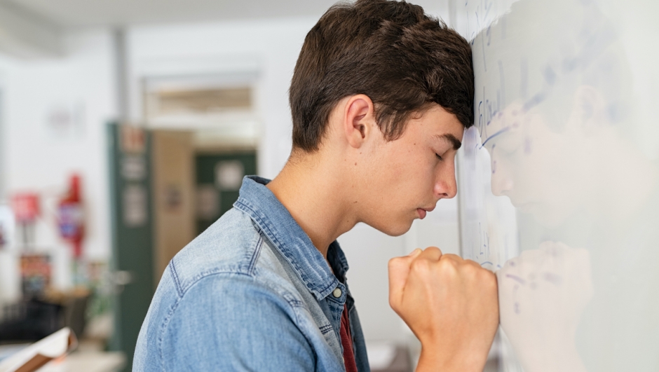 Disappointed university student leaning on whiteboard with closed eyes. Tired high school guy feeling failure in classroom. Side view of sad young college student leaning on whiteboard while trying to solve math problem.